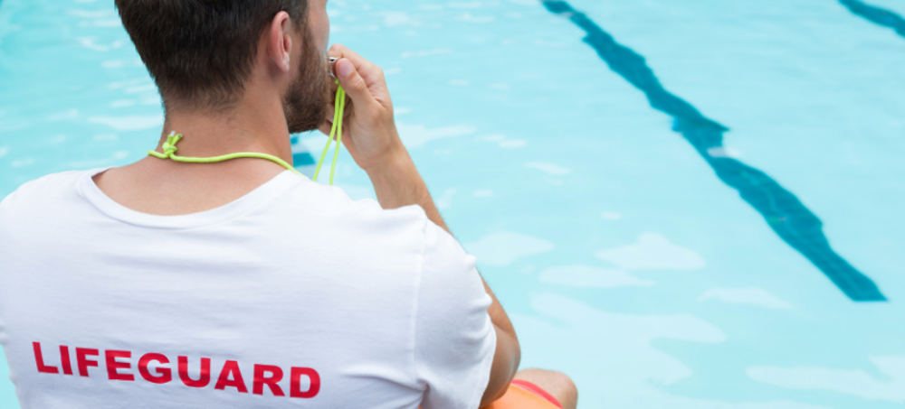 Man wearing lifeguard t-shirt sitting by pool, blowing a whistle