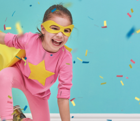 Girl dressed as superhero, wearing pink outfit and yellow cape and mask, with confetti tumbling around her