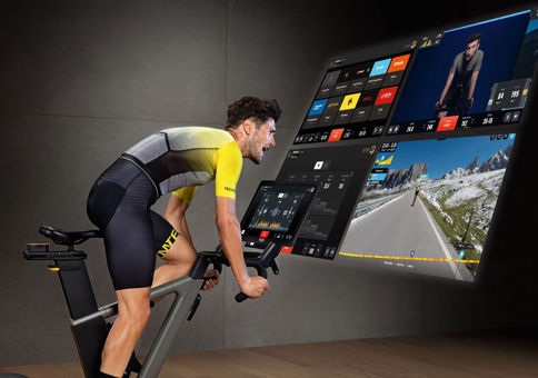 Side image of a man in lycra riding a stationary bike in a gym, while watching a large digital screen displaying a virtual route.