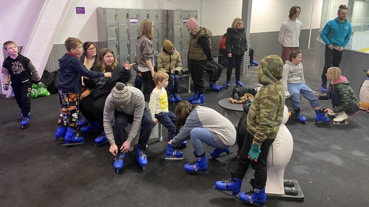 Group of adults and children sat on benches putting on blue ice skates at an indoor ice rink