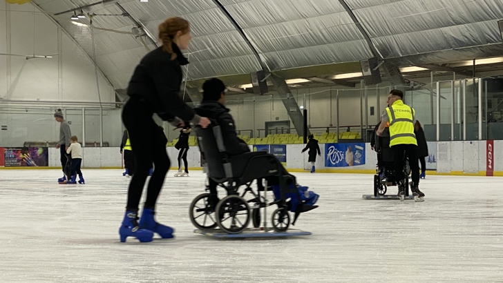Young woman on ice skates in an ice rink, pushing a person in a wheelchair along on the ice
