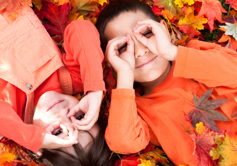 Two children in orange outfits laying in a pile of bright red and orange fallen leaves, making binoculars with their hands over their eyes.