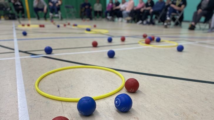 red and blue Boccia balls on the floor of a sports hall, next to a yellow hoop