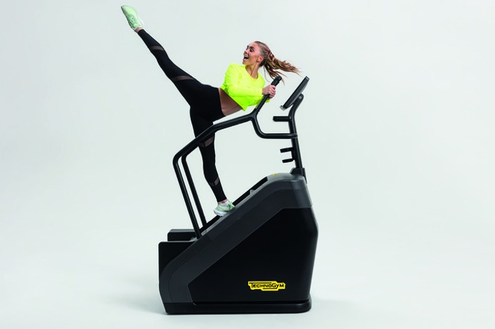 Energetic woman stood on a piece of stairclimber gym equipment, holding the handle and performing a high kick 