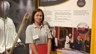 A woman with long, dark hair smiling in front of a London Olympics 2012 uniform on display. 