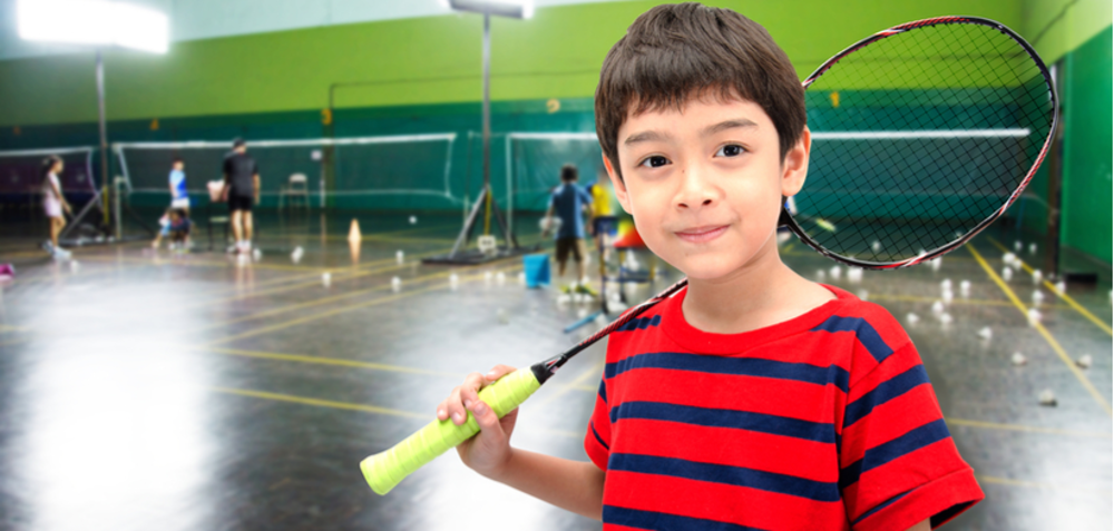 Young boy holding badminton racket in front of courts with other people playing