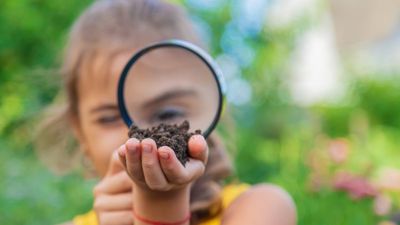 A young girl holds up a magnifying glass to her eye and looks at soil that she is holding in her hand. 