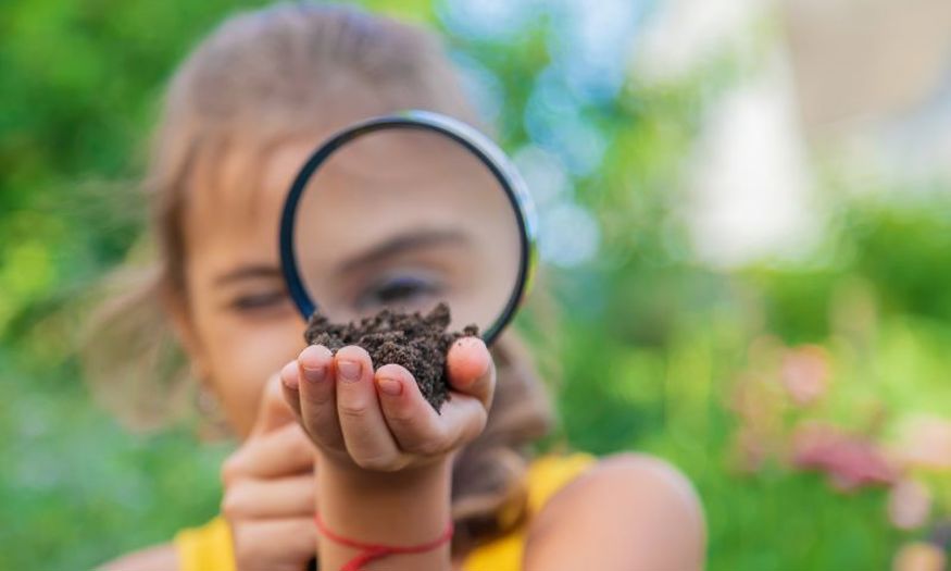 A young girl holds up a magnifying glass to her eye and looks at soil that she is holding in her hand. 