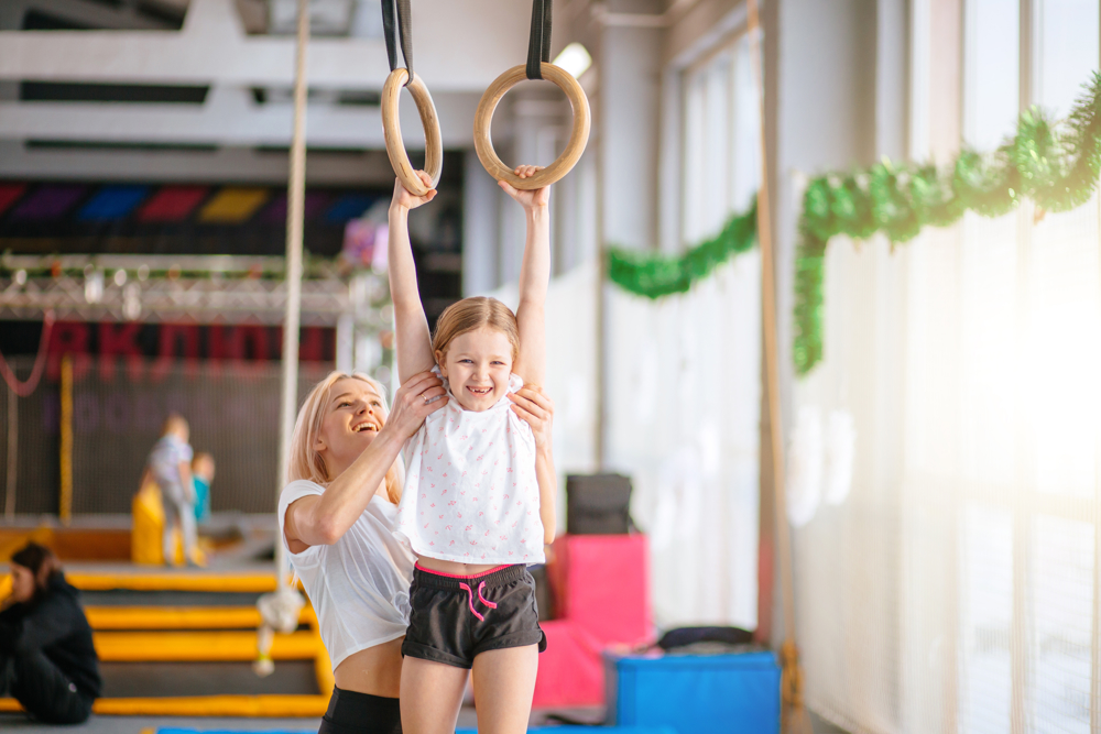 Young girl smiling being held up on gymnastics rings by a gymnastics teacher 